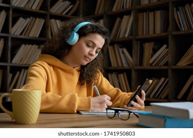 Latin teen girl wear headphones hold phone online learning in mobile app. Hispanic college student using smartphone watching video course, zoom calling making notes in workbook sit in library campus.