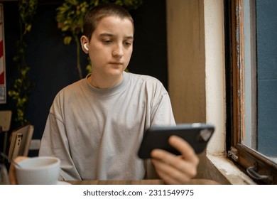 Latin transgender generation z student using mobile phone, digital tablet at the bar. He is using earphones for video call  - Powered by Shutterstock