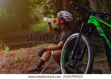 Latin woman cyclist sitting drinking water in the forest with her bicycle leaning against a tree