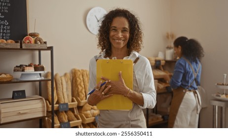 Latin woman holding clipboard smiling in bakery shop with colleague working in background - Powered by Shutterstock