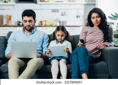 latin woman holding remote control while sitting on sofa near husband with laptop and daughter with digital tablet 