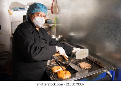latin woman looking at the camera while she is cooking a hamburger, wearing a mask, hat and gloves, in a small food shop in south america.