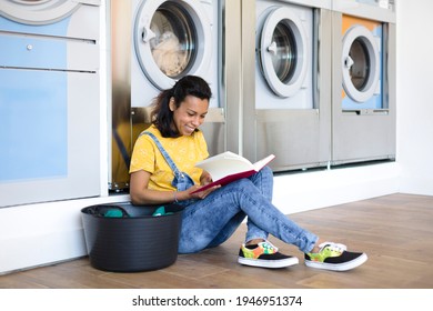 Latin woman reading a book sitting on the floor while waiting for clothes washing at the self-service laundry. She is smiling. - Powered by Shutterstock