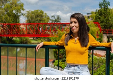 Latin woman sitting in a park with a lake, leaning on a railing with her arms open wearing a yellow sweater - Shutterstock ID 2219191179