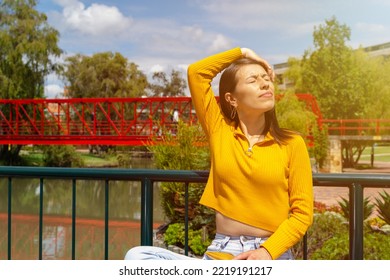 Latin woman sitting in a park with a lake, wearing a yellow sweater while touching her hair and enjoying a sunny day - Shutterstock ID 2219191217