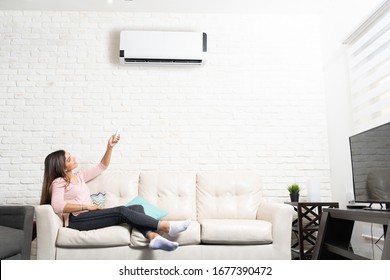 Latin woman turning on air conditioner unit using remote control while sitting on sofa at home - Shutterstock ID 1677390472