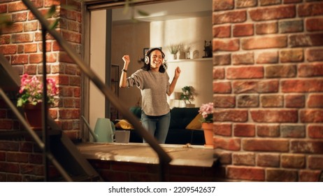 Latin Woman Wearing Headphones And Dancing In Apartment, Having Fun. Joyful Girl Feeling Happy And Carefree. Cheerful Female Is So Happy That You Want Join Her. Shot Throught Apartment Window.