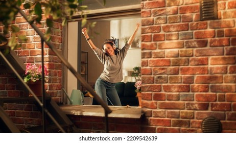 Latin Woman Wearing Headphones And Dancing In Her Apartment And Having Fun. Joyful Girl Feeling Happy And Carefree. Cheerful Female Is So Happy That You Want Join Her. Shot Throught Apartment Window.