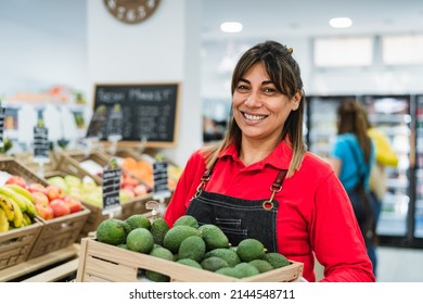 Latin woman working in supermarket holding a box containing fresh avocados - Shutterstock ID 2144548711