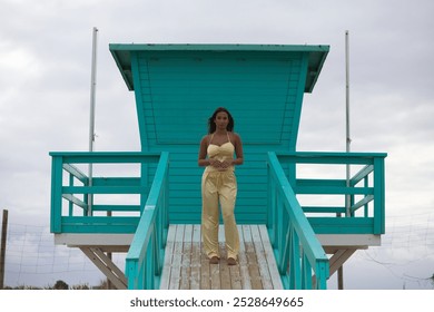 Latin woman, young and beautiful riding on the lifeguard stand on a beach in Cadiz, Spain. She is barefoot on the ramp and does different body postures. Blowing air and sea breeze - Powered by Shutterstock