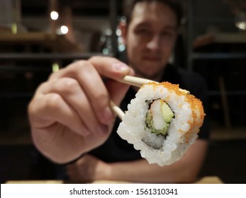 Latin young man with a surimi and avocado california roll maki picked with chopsticks. Focus on the maki. Eating sushi. Japanese restaurant.