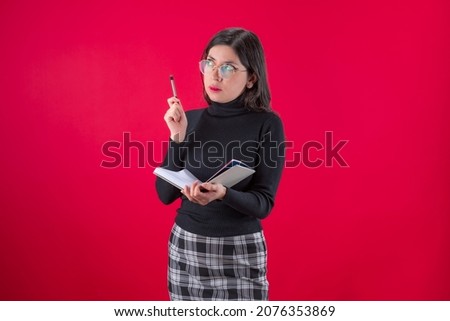 Latin young woman in black blouse and skirt poses doubtfully with a notebook and a pen in her hand on a red background.