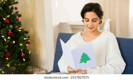 Latin young woman is smiling while reading a christmas card while sitting on a couch with a decorated christmas tree in the background - Powered by Shutterstock