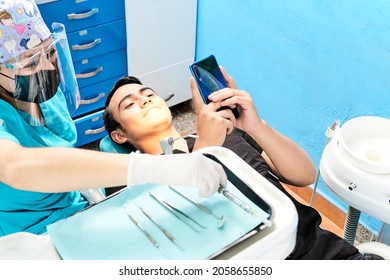 Latina Female Dentist Arranging Her Instruments While The Young Latino Male Patient Types A Message On His Smart Phone.