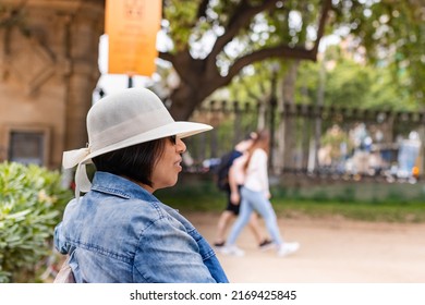 Latina Woman In Denim Shirt Sitting On A Bench In A Park In Barcelona (Spain).