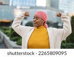 Latina woman, fighting breast cancer, wears a pink scarf, and clenches her arms as a survivor fighter