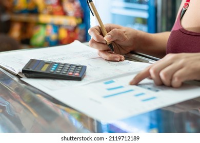 Latina Woman Pointing To Her Business Statistics Chart. Using Her Small Calculator And Cardboard Eco-friendly Pen On A Large Notebook. Doing Calculations On Her Grocery Store Sales.