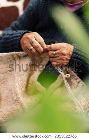 
Latina woman, senior adult, happy and focused, in the living room using knitting needles and sheep's wool, concept of traditional everyday life, traditional weaver, selective focus.