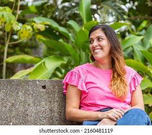 Latina Woman Sitting In A Bench In The Park Looking Away And Smiling