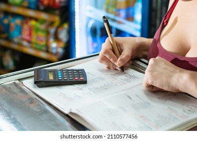 Latina Woman Writing With Her Eco-friendly Cardboard Pen On A Large Notebook And Doing Calculations On Her Calculator About Sales And Statistics In Her Grocery Store. On Top Of A Display Case