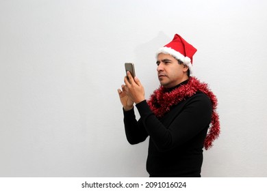 Latino Adult Man With Christmas Hat And Red Garland As Scarf Uses His Cell Phone To Call His Family With Happy Video Call And Excited To Celebrate Christmas
