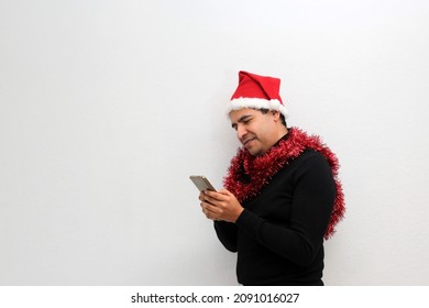 Latino Adult Man With Christmas Hat And Red Garland As Scarf Uses His Cell Phone To Call His Family With Happy Video Call And Excited To Celebrate Christmas
