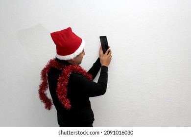 Latino Adult Man With Christmas Hat And Red Garland As Scarf Uses His Cell Phone To Call His Family With Happy Video Call And Excited To Celebrate Christmas
