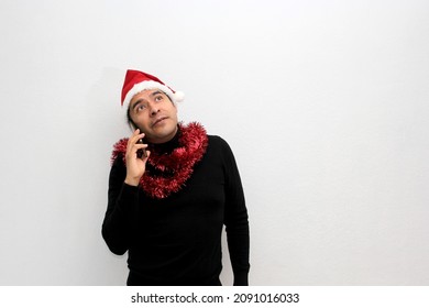 Latino Adult Man With Christmas Hat And Red Garland As Scarf Uses His Cell Phone To Call His Family With Happy Video Call And Excited To Celebrate Christmas
