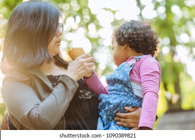 Latino American Mother And Dark Skinned Hispanic Toddler Daughter Eating Ice-cream During A Walk Outdoors.