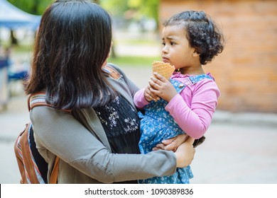 Latino American Mother And Dark Skinned Hispanic Toddler Daughter Eating Ice-cream During A Walk Outdoors.