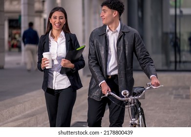 Latino Business Man And Woman Walking To Work. Young Latinos Walk Through The Streets Of The City On Bicycles.