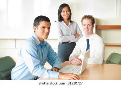 Latino Businessman Leader Sitting At Desk On Laptop In Conference Room, Meeting With Diverse Team Of Business People, Attractive Asian Woman And Caucasian Male Looking At Camera. Horizontal