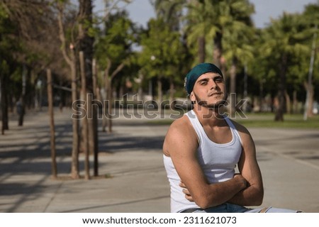 Latino and Hispanic boy, young, rebellious, with headscarf, arms crossed, smiling and looking defiantly at camera, sitting on a bench. Troubled concept, rebel, gangs, defiant.