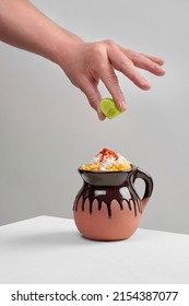 a latino, hispanic man squeezes a green lime over an Esquite, corn in a cup, a typical Mexican dish, in a clay cup with yellow corn, Mexican Huasteca cheese and chili powder on a white background