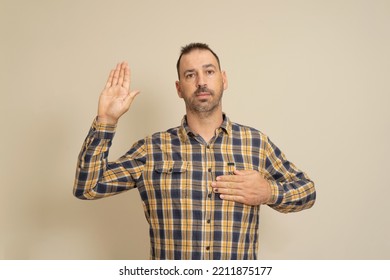 Latino Man In His Forties With A Beard Wearing A Plaid Shirt Smiling Swearing With His Hand On His Chest And Fingers Up, Making A Pledge Of Allegiance Oath.