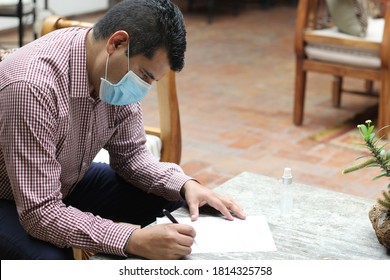 Latino Man With Protection Mask, Sitting In Waiting Room Reviewing And Signing Documents, New Normal Covid-19