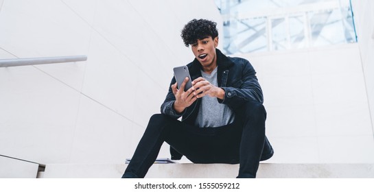 Latino Millennial Man Using Mobile Banking Application For Checking Account Balance, Shocked Hipster Guy In Trendy Wear Receive Message With Amazed News About Notification For Cellular Digital Phone