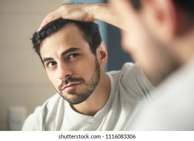 Latino person with beard grooming in bathroom at home. White metrosexual man worried for hair loss and looking at mirror his receding hairline. - Shutterstock ID 1116083306