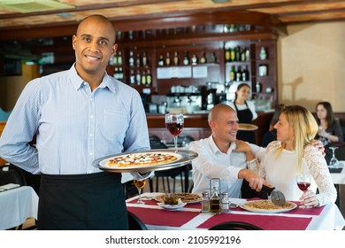 Latino Waiter With Tray Of Delicious Pizza On Tray Against The Background Of Restaurant Guests