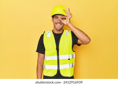 Latino worker in yellow vest and helmet excited keeping ok gesture on eye.
