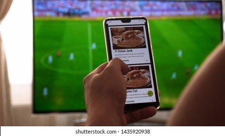 Latvia, Jurmala - 06 June 2019, Background Of The TV At Home (at The Bar), The Guy Orders Different Snacks, Pizza And Sweets Scrolling Through Internet Stores Using Mobile Phones. Concept: Sport TV.