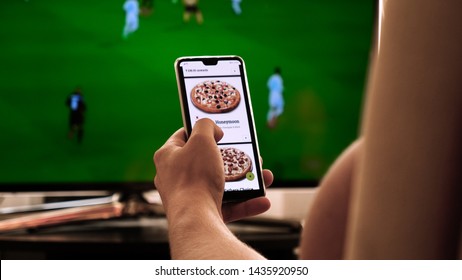 Latvia, Jurmala - 06 June 2019, Background Of The TV At Home (at The Bar), The Guy Orders Different Snacks, Pizza And Sweets Scrolling Through Internet Stores Using Mobile Phones. Concept: Sport TV.