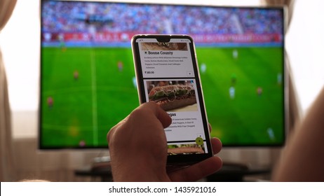 Latvia, Jurmala - 06 June 2019, Background Of The TV At Home (at The Bar), The Guy Orders Different Snacks, Pizza And Sweets Scrolling Through Internet Stores Using Mobile Phones. Concept: Sport TV.