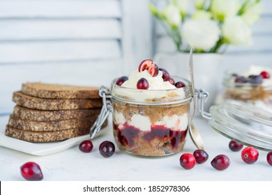 Latvian, scandinavian traditional rye whole grain bread layered dessert with whipped cream and cowberry jam served in glass jar on a white table with flowers and berries, copy space.