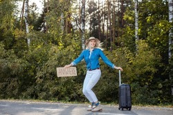 Laughing, Active, Cheerful Blond Female Hitchhiker In Dance Squat Position Hold Carton Board Anywhere With Luggage By The Road, Wait For Car Transportation To City Or Town Near Forest. Active Vacation