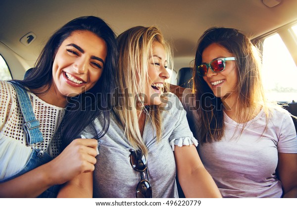 Laughing affectionate female friends riding in the\
back of a car on a road trip together smiling happily with\
enjoyment and pleasure at the\
camera