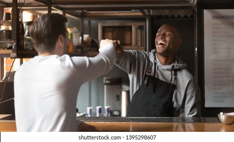 Laughing African American barista fists bumping with customer in cafe, excited bartender wearing apron standing behind bar counter, greeting friend or regular customer in coffeehouse - Powered by Shutterstock