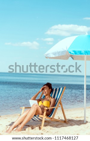 laughing african american woman relaxing on deck chair and using digital tablet under beach umbrella in front of sea 
