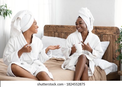 Laughing African American Young Women Having Spa Day At Home. Two Black Ladies In Bathrobes And Towels On Their Heads Having Conversation, Drinking Coffee, Bedroom Interior, Ladies Day Concept