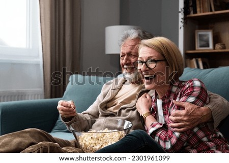 Laughing aged couple, man and woman watching tv, comedy show or movie and eating popcorn snack, sitting on cozy couch at home, mature family, man and woman enjoying free time, weekend together